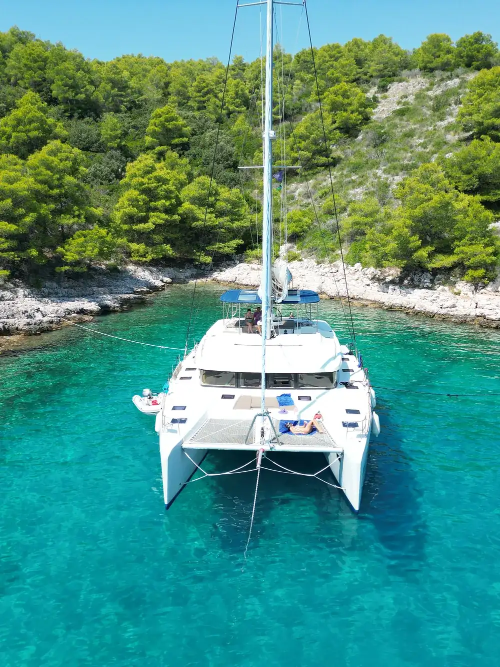 Crewed catamaran anchored bow-on in a pine-fringed Adriatic cove