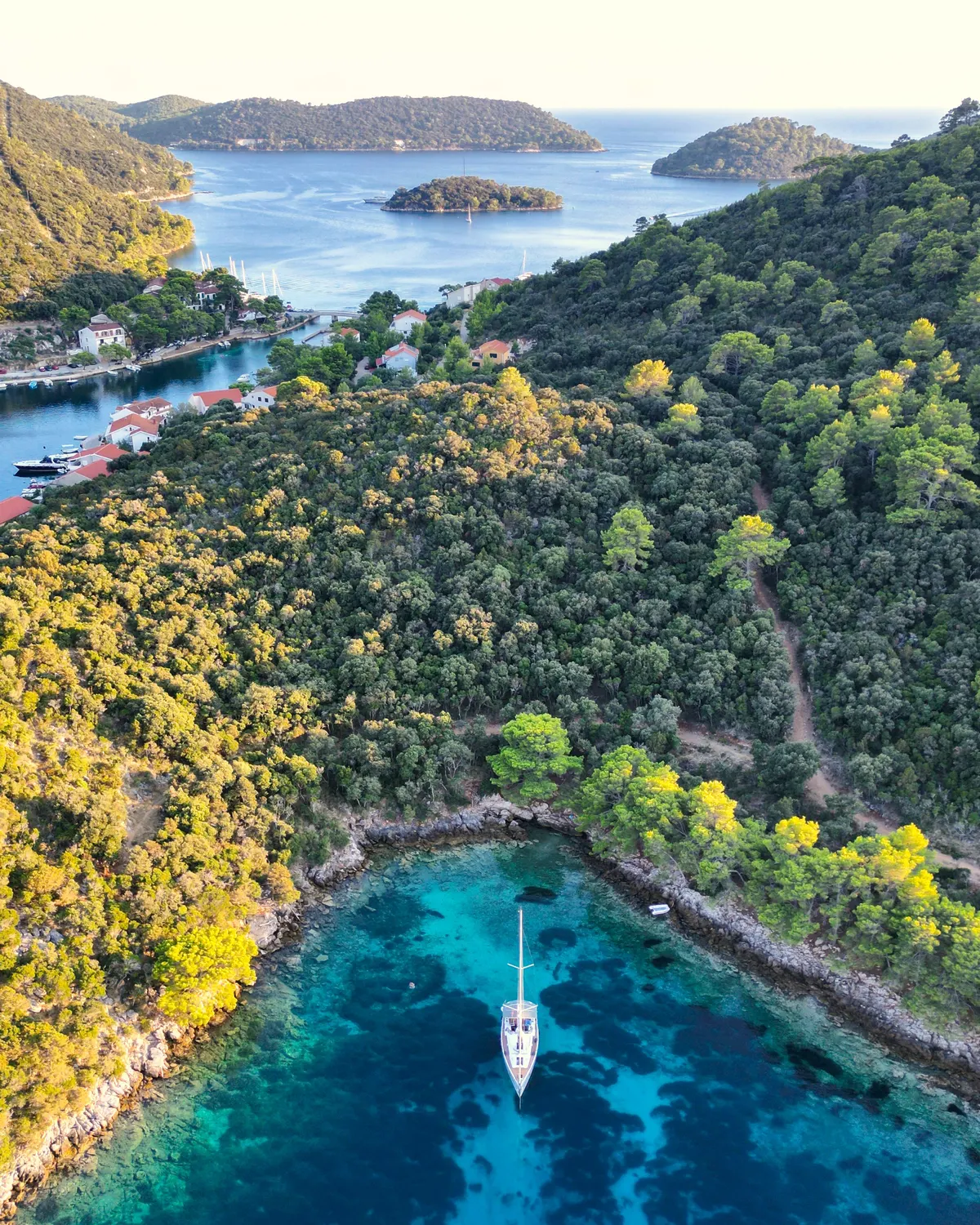 A sailboat at anchor alone in a clear turquoise cove ringed by pine forest, with small islands and a calm sea in the distance.